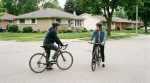 Two teenage riders stopped at the same suburban intersection, one with a foot down, the other still balanced.