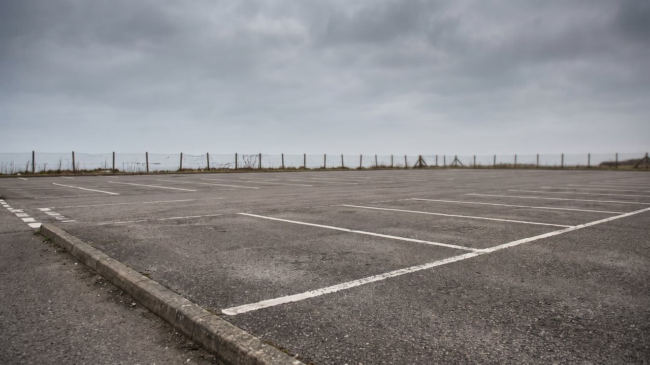 A wide empty asphalt parking lot early on a Sunday morning: two chalk lines drawn down one end, a few half tennis balls in a row, one machine on its stand and one adult sitting on a low wall.
